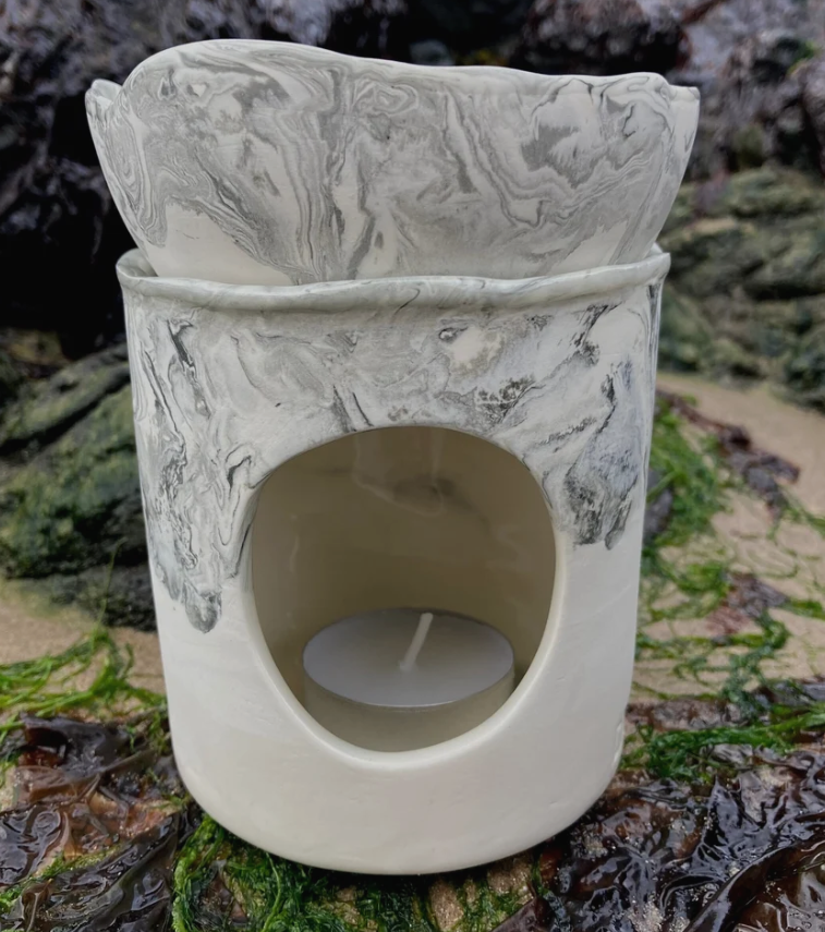A handmade ceramic candle warmer with a marbled gray and white glaze, featuring a top lid, a side cutout holding a white tea light candle, and a small matching dish on top, set against a natural rocky shoreline background.